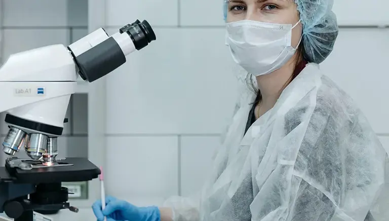 Seated masked scientist, in a lab, looks at us while sat at microscope